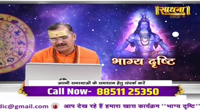 A man in orange sits behind a laptop, speaking on a television program called "Bhagya Drishti" from Sadhna Gold. Behind him, a celestial image of Shiva glows, and a phone number flashes on the screen.
