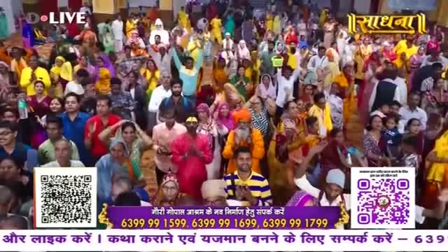 A large crowd, many in colorful clothing, are gathered, with their arms raised. The word "Sadhna" is visible at the top, and there's a QR code displayed on the screen.
