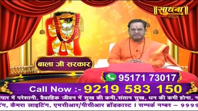A man in orange robes sits behind a red table, facing the camera on a Sadhna show from India. A phone number flashes on the screen, inviting viewers to call for help.
