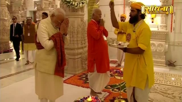 The man in the cream-colored outfit stands with hands clasped, head bowed in prayer. A priest in yellow raises a tray, while another man in red stands nearby.
