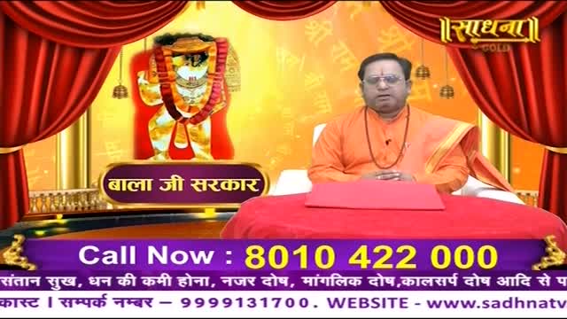 A man in orange robes sits behind a red table, speaking into a camera on the Sadhna channel. Behind him, a colorful image of a deity is displayed, and a phone number flashes on the screen.
