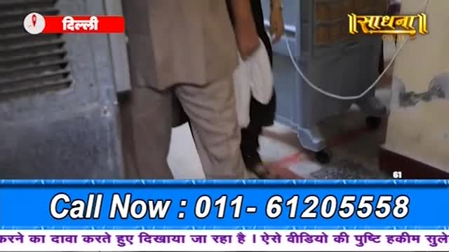 A person in a light suit holds a white cloth, walking past a wall. The Sadhna logo and a call-in number are displayed on the screen.
