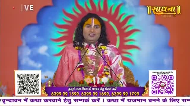 A man in a pink and patterned shirt speaks into two microphones, a backdrop of a sun-like design behind him. The Sadhna Gold logo is visible, and QR codes and phone numbers are displayed at the bottom of the screen.
