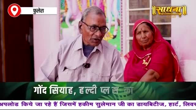 An older man with glasses speaks, a small microphone clipped to his shirt. Beside him, a woman in a red headscarf listens intently, both seated in front of a backdrop with text in Hindi.

