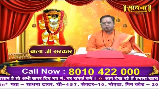 A man in an orange robe sits at a red table, speaking on a Sadhna Gold broadcast. Behind him, a framed deity image and ornate curtains create a backdrop for the show.
