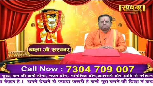 A man in an orange robe sits at a red table, facing the camera. Behind him, a golden backdrop features an image of a deity and the Sadhna logo.
