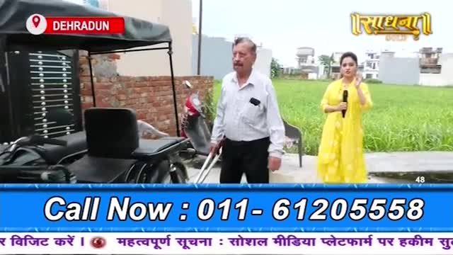 A man in a white shirt stands beside a modified wheelchair, holding a cane. A woman in a yellow dress gestures with a microphone in front of a grassy field, as the Sadhna logo is visible in the top right.
