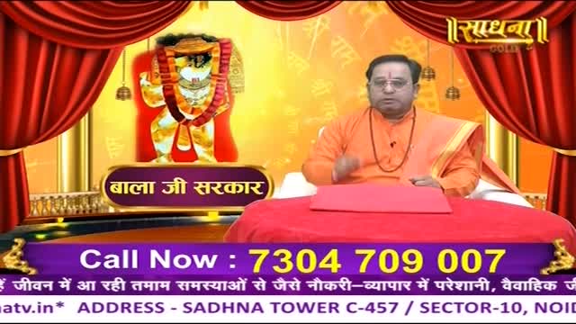 A man in orange robes sits behind a red table, gesturing with his hands. Behind him, a golden backdrop features an image of a deity, and the Sadhna Gold logo is visible.
