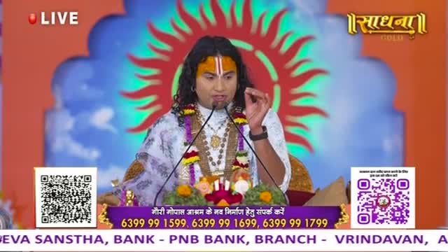 A man with a painted forehead and long dark hair speaks into two microphones, gesturing with his right hand.  Behind him, a bright sunburst design and the Sadhna Gold logo fill the backdrop.
