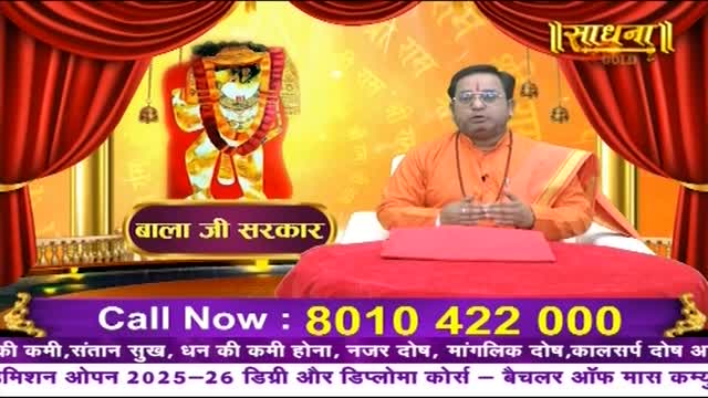 A man in orange robes sits behind a red table, gesturing with his hands. Behind him, a deity's image is displayed, and the Sadhna logo is visible at the top.
