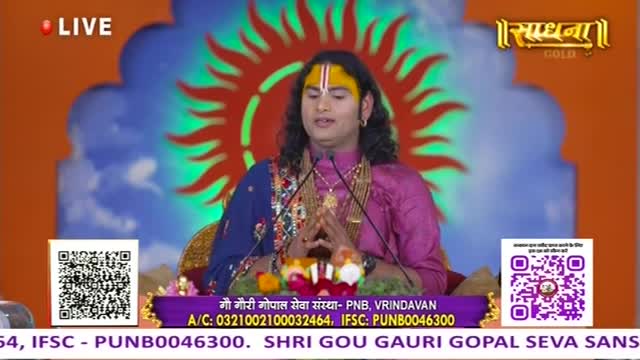 A man in a pink and blue outfit sits before microphones, speaking with hands clasped. The Sadhna Gold logo and a sun design are visible behind him.
