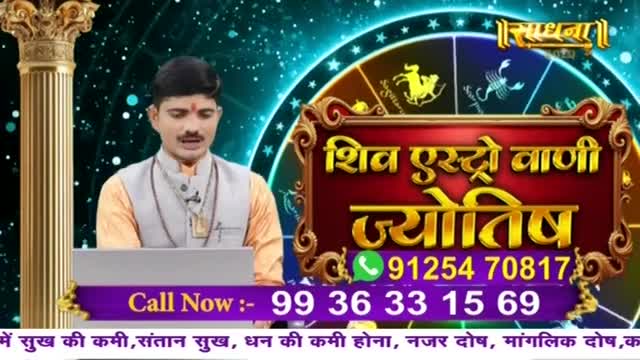 A man in a waistcoat speaks from behind a small desk, the backdrop displaying a swirling cosmic design and Hindi text. The Sadhna logo and phone numbers for astrological services are prominently featured on the screen.
