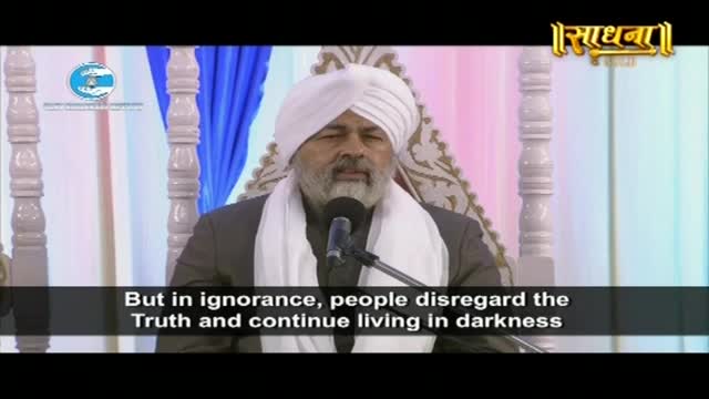 A man in a white turban speaks into a microphone. He wears a dark suit and a white scarf.