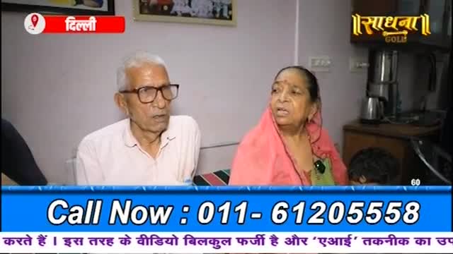 An older man and woman sit side-by-side, their faces turned towards the camera. The woman wears a bright pink sari, and a logo for "Sadhna Gold" is visible in the background.