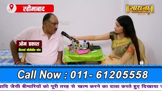A man in a pink shirt speaks into a microphone held by a woman in a sari. Bottles of medicine and other items are arranged on a green cloth-covered table between them.