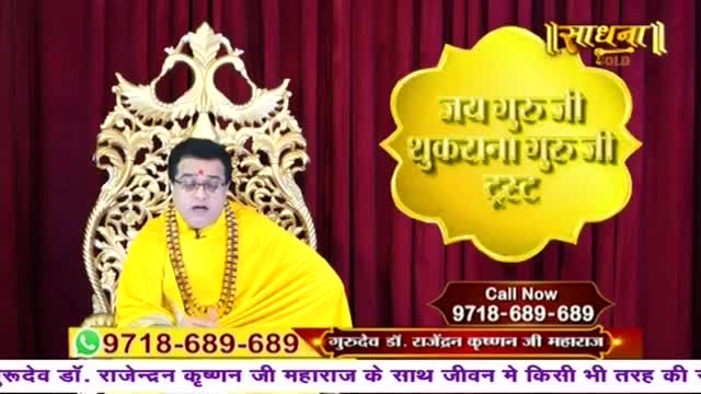A man in a bright yellow robe sits on an ornate chair, speaking into a microphone. A golden cloud-shaped graphic with Hindi text hovers to his right, and a phone number is displayed below.