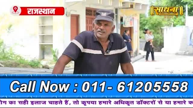 A man in a dark blue shirt with white stripes stands talking, his expression earnest. Behind him, a woman carries a child down a dusty street in Rajasthan.