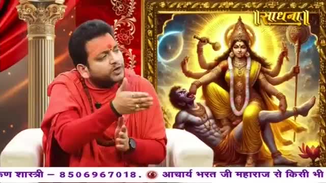 A man in red robes gestures emphatically while speaking. Behind him, a vibrant painting depicts a multi-armed goddess triumphing over a figure.