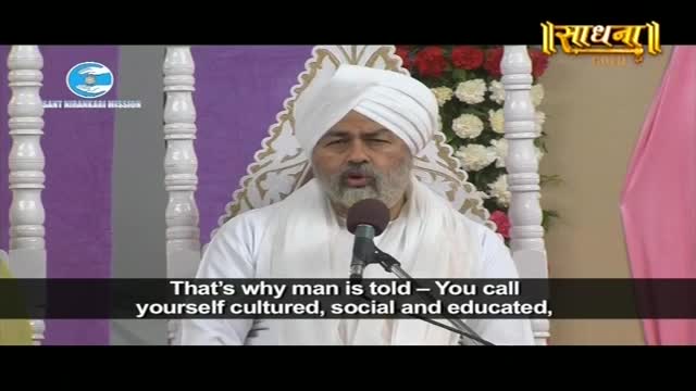 A man in a white turban speaks into a microphone, his words appearing as subtitles. He is seated before a backdrop of purple fabric and white pillars, with floral arrangements to his right.