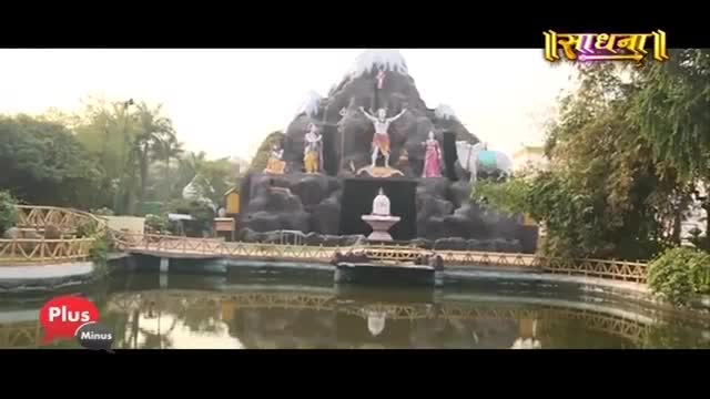 A large, rocky structure with statues of deities stands behind a calm pond. A wooden bridge arches over the water, leading towards the monument.
