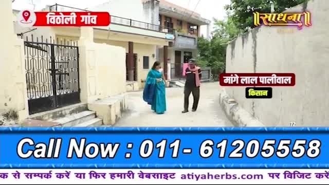 A woman in a bright blue sari walks away from a gate, while a man in a dark shirt and trousers, with a pink scarf around his neck, walks towards her. The scene is set in a village in India, with modest houses lining a narrow lane.