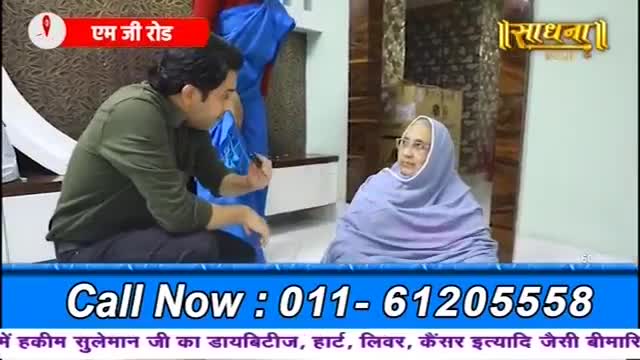 A man in a green shirt leans forward, holding a pen, while an older woman in a purple headscarf sits opposite him. The Sadhna logo is visible in the background.