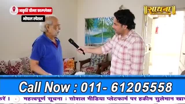 A man in a blue shirt is being interviewed by another man holding a microphone. The interview appears to be for a program called Sadhna, possibly from Bhopal.