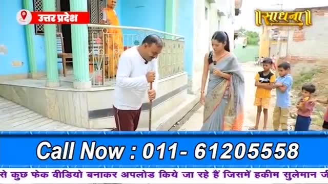 A man in a white shirt and maroon pants kneels, sweeping the ground with a long stick. A woman in a grey sari stands nearby, watching him.