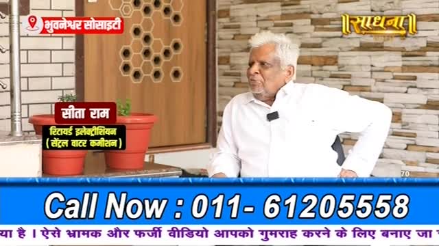 A man with white hair sits and speaks, a microphone clipped to his shirt. Two red planters sit in front of him, one with a small plant.