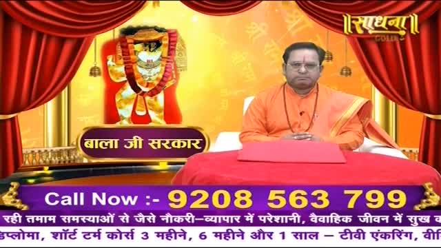 A man in an orange robe sits before a vibrant backdrop, a framed image of a deity behind him.  A phone number flashes across the bottom of the screen, inviting calls for solutions to life's problems.