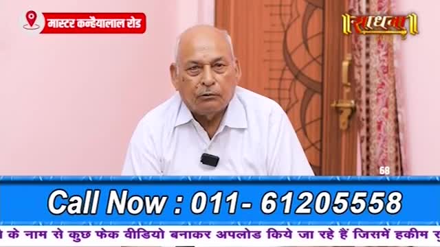 An older man with a serious expression sits in front of a pink wall. A channel logo, "Sadhna," appears in the upper right corner. Below him, a blue banner displays a phone number and text in Hindi.