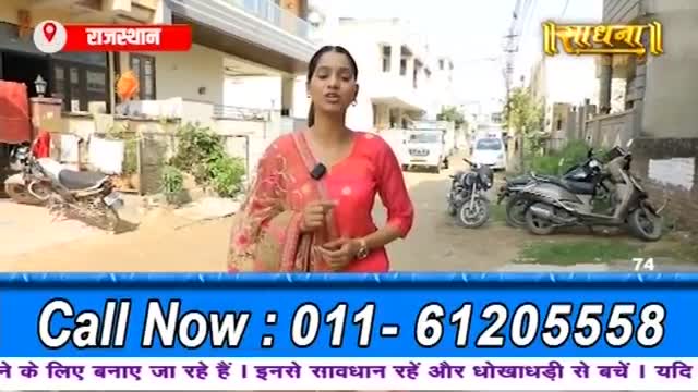 A woman in a bright pink outfit speaks into a microphone on a dusty street in India. Several motorcycles and scooters are parked along the side of the road.