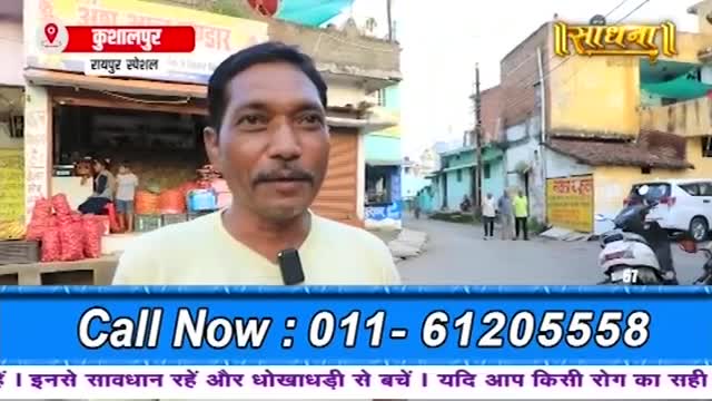 A man stands in front of a shop, speaking into a microphone. Behind him, a street in India bustles with a few people and parked vehicles.
