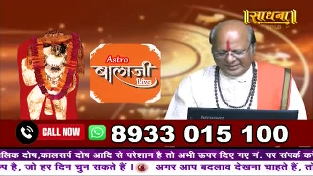 A man in traditional Indian attire, likely a pundit, looks down at a laptop. To his left, a colorful idol of Hanuman stands adorned with garlands. A phone number flashes across the screen, inviting calls.