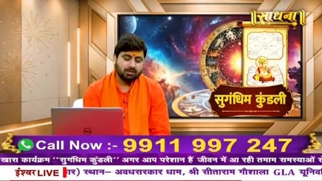 A man in an orange robe speaks into a microphone, his gaze directed downwards. Behind him, a screen displays a vibrant astrological chart and the words "Sugandhim Kundali."