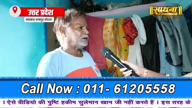 A man with a grey beard is speaking into a microphone held by a person off-camera. Clothes hang on a rack behind him.