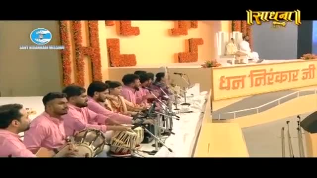 A group of men in pink shirts sit in a row, their hands poised over tabla drums. A large banner with Hindi script hangs behind them, and a man in white sits on a throne at the back of the stage. A group of men in pink shirts sit in a row, their hands poised over tabla drums. A large banner with Hindi script hangs behind them, and a man in white sits on a throne at the back of the stage.