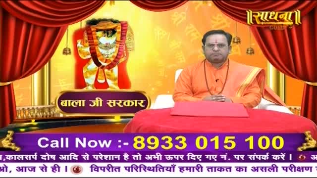 A man in an orange robe sits behind a red table, his hands clasped. A vibrant image of a deity, likely from India, hangs on the wall behind him, framed by heavy red curtains.