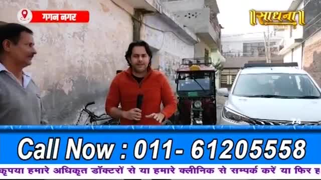 A man in an orange jacket speaks into a microphone, gesturing with his hands. Beside him, a small, red auto-rickshaw is parked next to a white car.