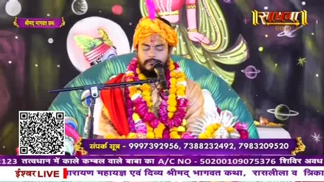 A man in a yellow turban and floral garlands speaks into a microphone. He's seated before a backdrop depicting planets and a stylized figure.