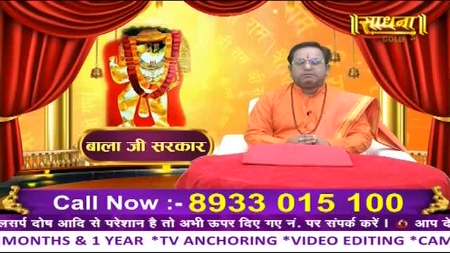 A man in an orange robe sits behind a red table, his hands clasped. Behind him, a portrait of a deity hangs, framed by red curtains.