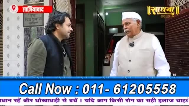 An older man in a white cap and vest speaks animatedly with a younger man wearing a dark vest over a green shirt. The conversation happens outside a building, with a car visible behind the older man.