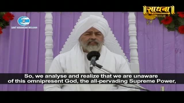 A man in a white turban and robes speaks into a microphone, his beard framing his face. The backdrop is a soft purple curtain, adorned with white pillars and a few red flowers.