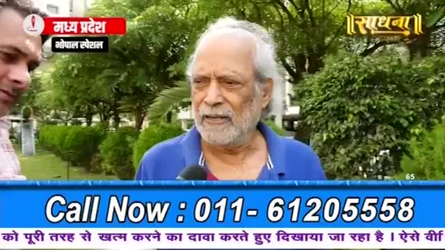 An older man with white hair and a beard speaks into a microphone, his blue shirt collar visible. Behind him, lush green trees and bushes form a natural backdrop.