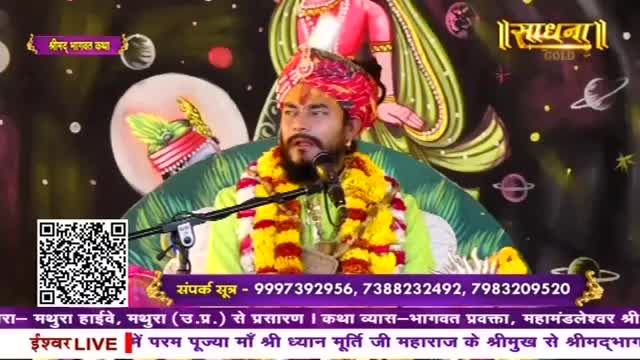 A man in a vibrant turban and floral garlands speaks into a microphone. He sits before a backdrop depicting celestial bodies and figures.