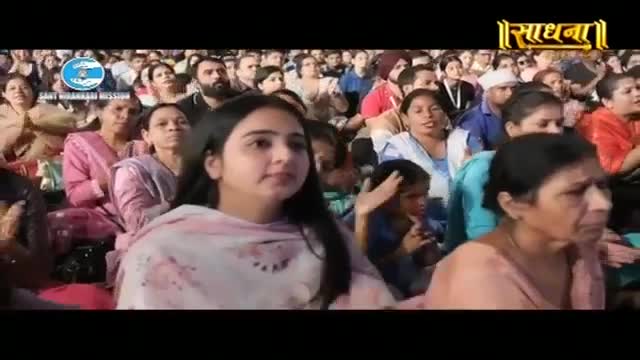 A crowd of people sits attentively, their faces turned towards something unseen. A young woman in a pink and white salwar kameez is in the foreground, her gaze fixed ahead.