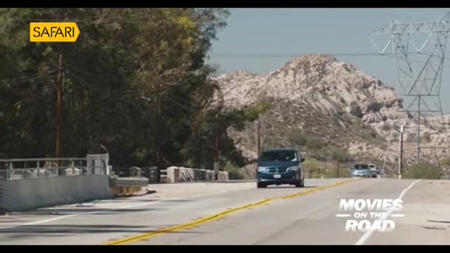 A dark blue SUV cruises down a sunny road, heading toward a mountainous backdrop. The Safari TV logo is visible in the upper left corner, hinting at a movie scene.
