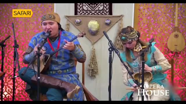 On a stage, a man in a blue embroidered tunic strums a long-necked instrument and sings into a microphone. Beside him, a woman in a matching outfit bows a stringed instrument, the Opera House logo visible in the corner.
