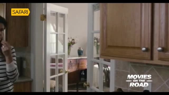 A woman in a striped shirt stands in a kitchen, a Safari TV logo visible in the corner. Through a set of French doors, a table and vase of flowers are visible.
