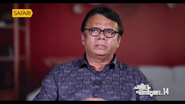 The man in glasses, likely a host on Safari TV, is speaking, his mouth moving as he looks slightly off-camera. A red backdrop and the show's title card suggest a studio setting in India.
